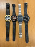 Four wristwatches displayed flat on a wooden surface: two black digital sports watches with rubber straps, one gold-tone plastic digital sports watch, and one silver-tone metal bracelet calculator watch by Elex missing its back cover.
