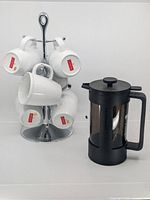 Bodum French press with six white Bodum cups on chrome stand, cups show red logo on bottom, stand has central handle and holds cups by handles.