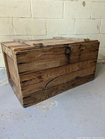 View of the wooden crate showing solid wood planks, metal latch on front, and visible crack on bottom front plank.