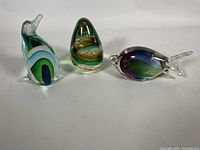 Three colorful blown glass sculptures/paperweights on white background: penguin, teardrop, fish shapes
