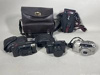 Photo showing the three cameras arranged on a white background with two camera carrying cases behind them: one large dark brown leather bag and a smaller black soft case with red accents.