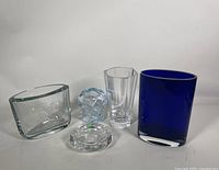 Five art glass pieces shown side-by-side on a white background. From left to right: clear low bowl, small clear glass animal figure, clear cut glass vase, large cobalt blue vase, and baby blue Orrefors signed vase.