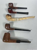 Five vintage tobacco pipes displayed together on a surface, including briar wood and one carved meerschaum pipe with visible wear and a broken stem.