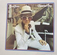 Elton John Greatest Hits vinyl album cover showing Elton John wearing white suit, large white hat, and large pink glasses, sitting in front of piano.