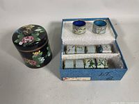 Photo showing the antique Chinese cloisonné tobacco humidor with carved brass foo-dog finial next to the open blue original box containing 10 cloisonné napkin rings displayed with foam padding.