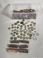 Plastic bin partially full of mixed coins plus laid out rows of assorted coins including Canadian pennies, pesos, and euros, with five rolls of Canadian pennies.