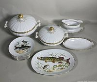 Full view of all porcelain dishes including fish motif plates, two covered serving dishes with lids, gravy boat with underplate and oval serving dishes.