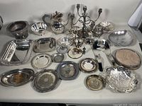 Overall view of the entire collection displayed on a white surface, showing various silver plated trays, bowls, candelabra, teapot, and other serving pieces in vintage condition.