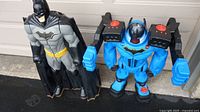 Photo showing two Batman toys side by side: one black with yellow belt and cape, about 33 inches tall; the other blue robotic-style with large arms, about 23 inches tall.