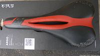 Top-down photo of Fizik Argo Vento R3 bike saddle on original black backing. Shows black and red textured surface, central cut-out, and branding.