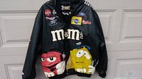 Front view of black genuine leather NASCAR jacket with large embroidered M&M's characters and sponsor patches.