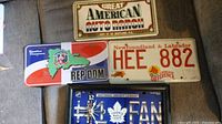 Four metal license plates shown on a fabric surface. Plates include Great American Auto Ranch, REP DOM, Newfoundland & Labrador HEE 882, and Toronto Maple Leafs FAN with clock.
