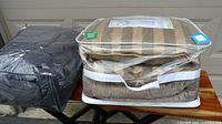 Grey comforter and beige comforter in plastic bag, plus two pillows on table.