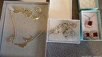 Three sets of costume jewelry displayed in boxes: gold-tone necklace with beads, silver-tone chain with cross charms, and silver-tone jewelry with red stones and rhinestones.