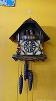 Full frontal view of the vintage Cuendet cuckoo clock mounted on yellow wall, showing chalet roof, clock face with Roman numerals, figurines, and pinecone weights.
