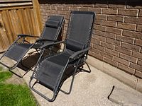 Two black outdoor folding lounge chairs with mesh fabric reclining on concrete patio next to brick wall.
