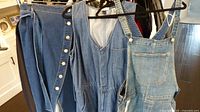 Three denim items hanging side-by-side: skirt, sleeveless dress, and overalls.
