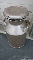 Full view of the vintage metal milk can showing the cylindrical body, two side handles, and the lid on top.