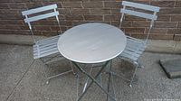 Full view of metal round bistro table and two chairs set on concrete ground against brick wall, all painted silver, foldable and clean condition.
