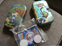 Two Pokémon-themed metal tins with colorful artwork and a bag of Pokémon trading cards in front.