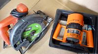 Two power tools: Black & Decker circular skill saw with orange handle and green FAB blade, Black & Decker orange router in a black storage container.