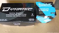 Photo showing closed black box of Dynamic safety glasses labeled 12 pairs next to blue-black box of Benehal N95 Particulate Respirator masks.