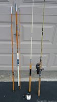 Full view of three fishing rods standing upright, showing orange, white, and olive-green rods with white and cork handles and mounted reels.