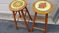 Two wooden stools with twisted legs and vegetable artwork on seats, shown from side angle.