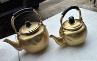 Two golden aluminum kettles side by side, showing size difference and classic round shape with spout, black handle, and knob lid.