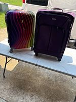 Two large suitcases side by side on a white folding table outside: left Dejuno hard shell multi-color, right purple American Tourister soft case