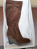 Left side view of one brown suede knee-high wedge boot inside white box showing the upper shaft and part of the wedge heel