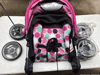 Disney baby stroller folded with pink, white, black, and green polka dot fabric seat, surrounded by detached stroller wheels on a white table.