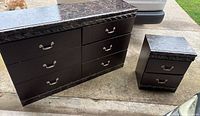 Photo showing the dresser and night stand set side by side on a driveway surface. The tops of both pieces have black marble and the furniture shows scratches and paint chips.