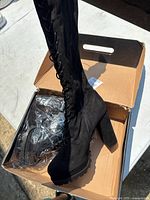 Black lace-up boots displayed in an open cardboard shoe box on a white table outdoors, showing side and front views of the boots with clear lighting highlights the texture and details.