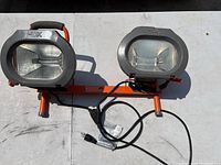 Front view of the HDX dual work light unit showing two rectangular light fixtures on an orange frame with power cord and handle.