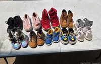 Photo showing 12 pairs of kids shoes arranged on a white table in two rows. Various colors and styles visible including black, pink, red, brown, iridescent, blue, yellow, and white.