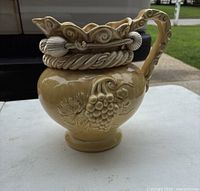 Front angled view of large beige ceramic pitcher with grape and leaf embossing, twisted rope and seashell design on top rim and handle.