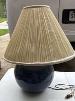 Photo showing the lamp's light blue round ceramic base and the beige pleated lampshade from the front with cord and switch visible on the table.