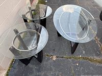 Set of one oval coffee table and two round end tables with glass tops and wooden bases on outdoor pavement