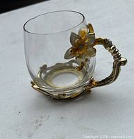 Glass mug with gold-tone decorative handle featuring gold enamel flower and curved design, base with matching metalwork, small chip on rim visible.