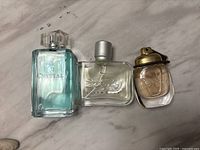 Three used fragrance bottles on marble surface: Watermark Crystal (light blue), Lacoste (clear), and an unlabeled small round bottle with gold cap.