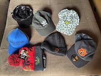 Seven hats displayed on a sofa, showing variety including Adidas cap, Auburn cap, leopard print cap with 'Babe' embroidery, knit beanies, and others