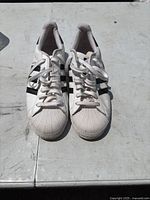 Pair of white Adidas sneakers with black side stripes and shell toe design on a concrete surface, viewed from front showing the laces and toe caps.