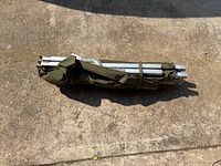 Folded metal cot frame wrapped in green canvas secured with straps, laid on concrete surface.