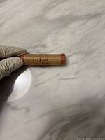 Roll of pennies labeled '1964' showing side and end of paper roll held on white marble surface by gloved hand.