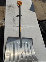 Snow shovel laid flat on a table showing the full length from orange grip handle to metal scoop, photo taken to display condition and overall size.