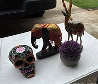Photo showing four decorative items: wooden elephant, painted antelope, ceramic skull, and small faux plant on a white table surface.