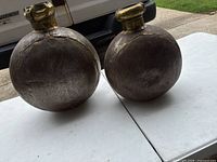 Photo showing two round vintage Indian metal water jugs with brass tops on white table outdoors.