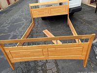 Queen size wooden bed frame parts laid out on asphalt, including headboard, footboard, side rails, and wooden slats.