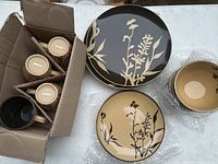 Overview of set components including stacked plates, bowls, and boxed cups showing ceramic with beige and black floral shadow design